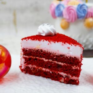 A vibrant red velvet cake slice with frosting, surrounded by festive colorful decorations.